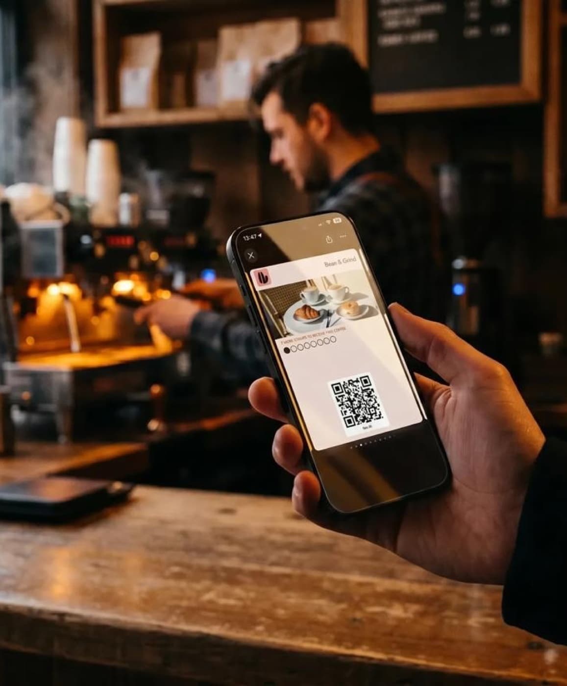 A customer at the café counter holds their phone showing their coffee loyalty card, with the barista and espresso machine behind